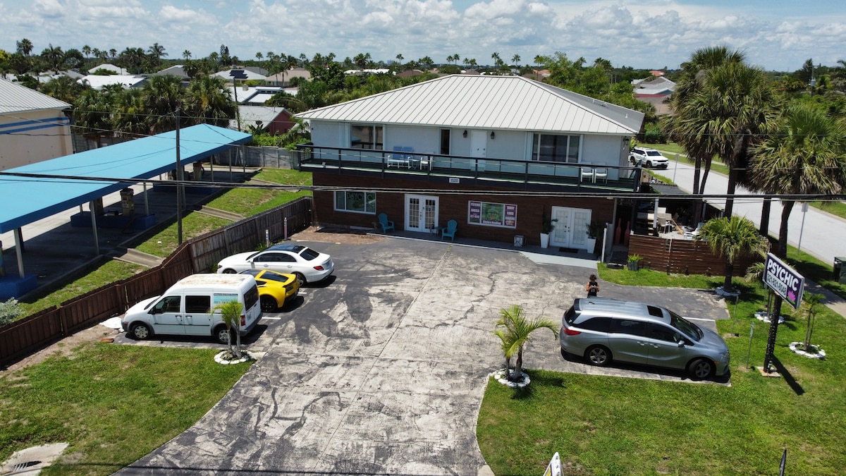 An aerial view of the property reveals a two-story building surrounded by palm trees and grassy areas. A spacious parking lot accommodates several vehicles, including a white van and a yellow car. Covered parking spaces are present on the left.