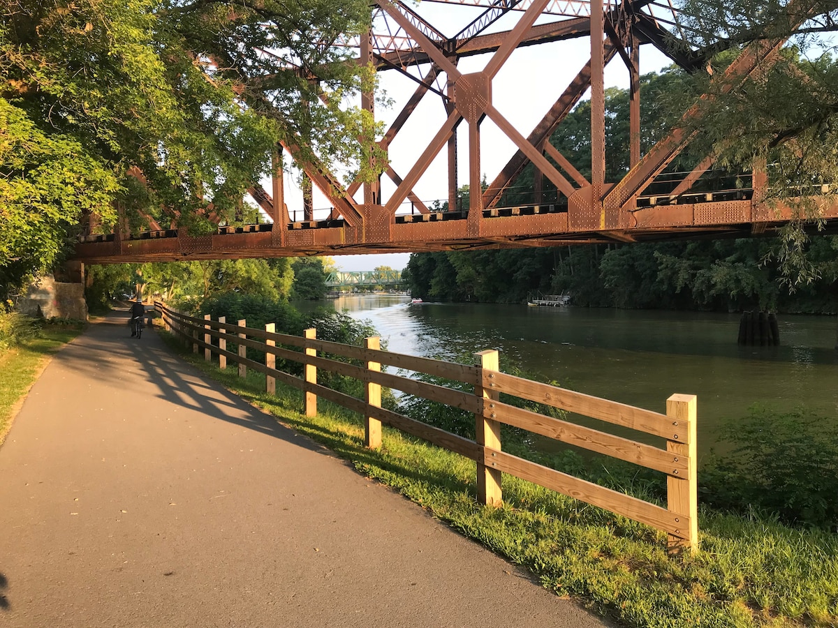 A steel bridge spans over a calm river, surrounded by lush greenery. A paved path is visible in the foreground, bordered by a wooden fence. The serene water reflects the colors of the trees and sky, creating a peaceful natural setting.