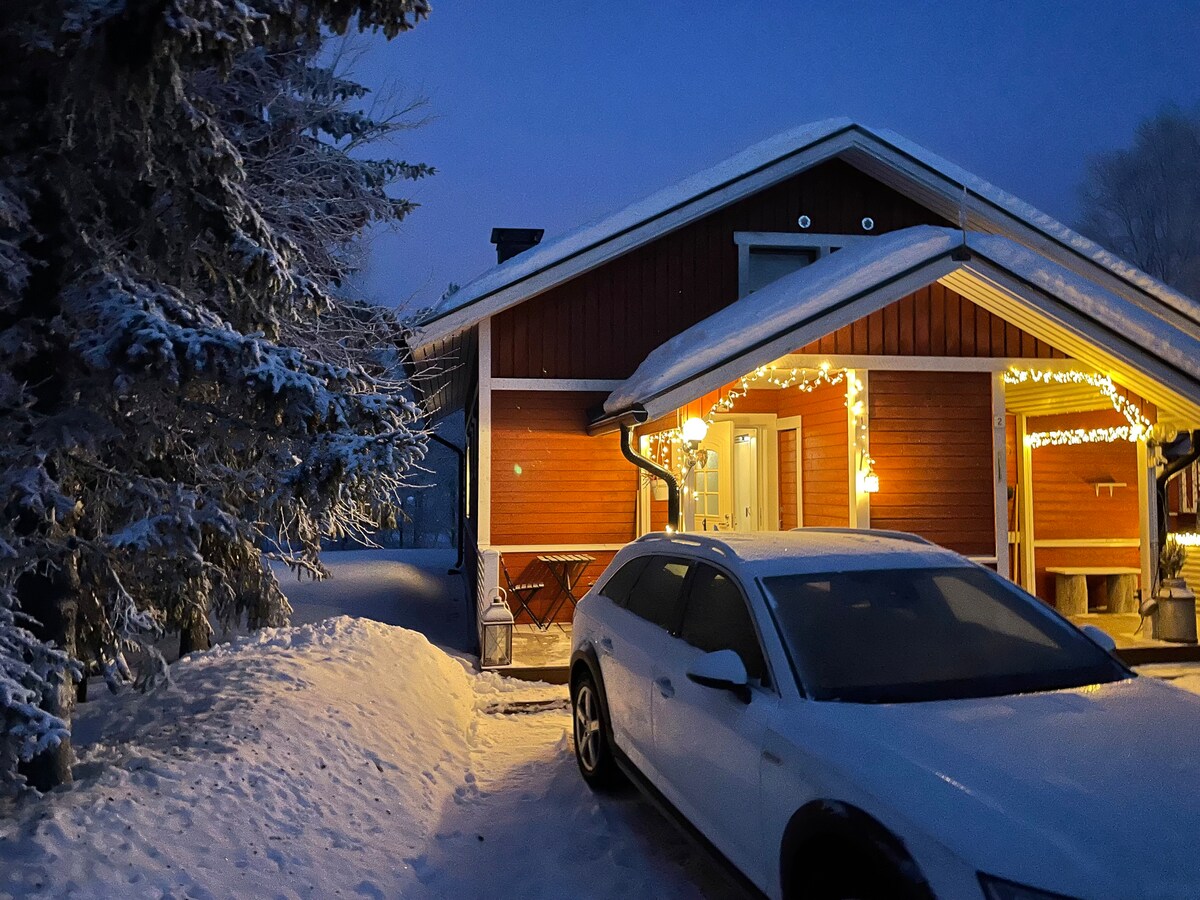 A cozy wooden house is displayed in the evening with snow covering the ground. Warm lights outline the entrance, enhancing the welcoming exterior. A parked white vehicle is visible, alongside snow-covered bushes, creating a serene winter atmosphere.