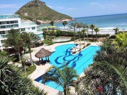 A serene pool area is framed by palm trees, featuring a large pool with a winding design. A thatched roof structure provides shaded seating. In the background, the beach and a rocky hill are visible under a clear blue sky.