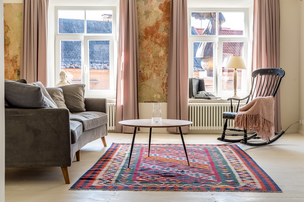 A living area features a comfortable gray sofa and a rocking chair, accompanied by a round coffee table. The space is anchored by a colorful patterned rug. Two large windows with pale curtains provide natural light, revealing a glimpse of the building's charming exterior.