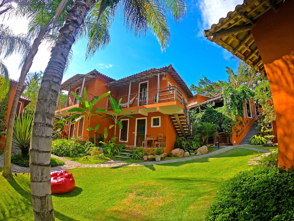 The exterior of the property is visible, showcasing a vibrant orange building surrounded by lush greenery. Palm trees and various plants frame the scene, while a well-maintained lawn offers a comfortable outdoor area. Balconies are seen on the upper level, enhancing the inviting atmosphere.