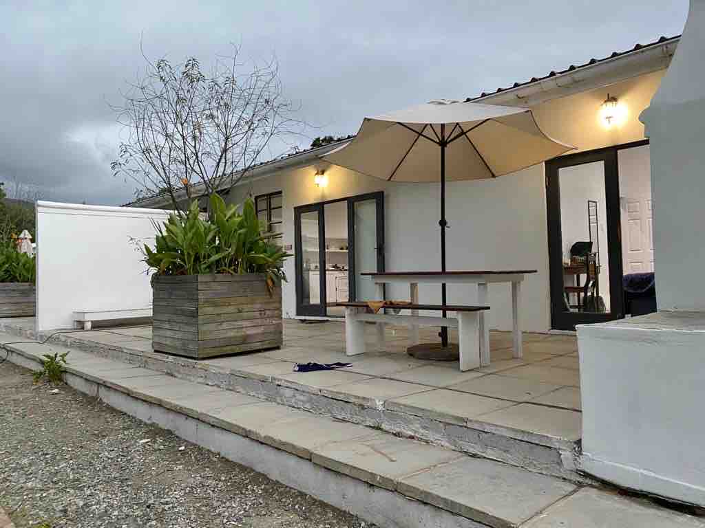 An outdoor patio area is featured, with a large umbrella providing shade over a wooden table and benches. Potted plants are arranged in a wooden planter. The building's exterior is painted white and includes large glass doors that invite natural light inside.