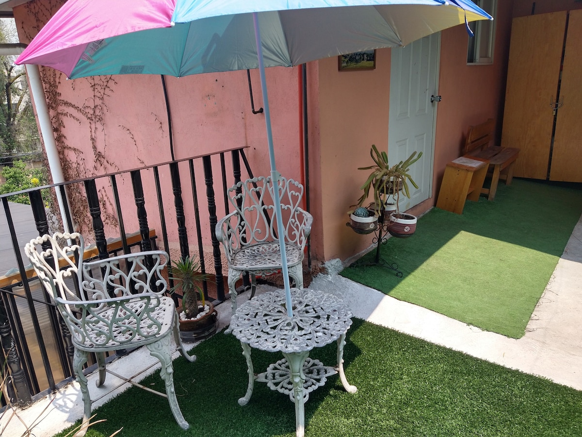 An outdoor patio area features ornate metal furniture consisting of a round table and two chairs, shaded by a colorful umbrella. Lush green artificial grass surrounds the seating, and potted plants are positioned nearby, enhancing the inviting nature of the space.