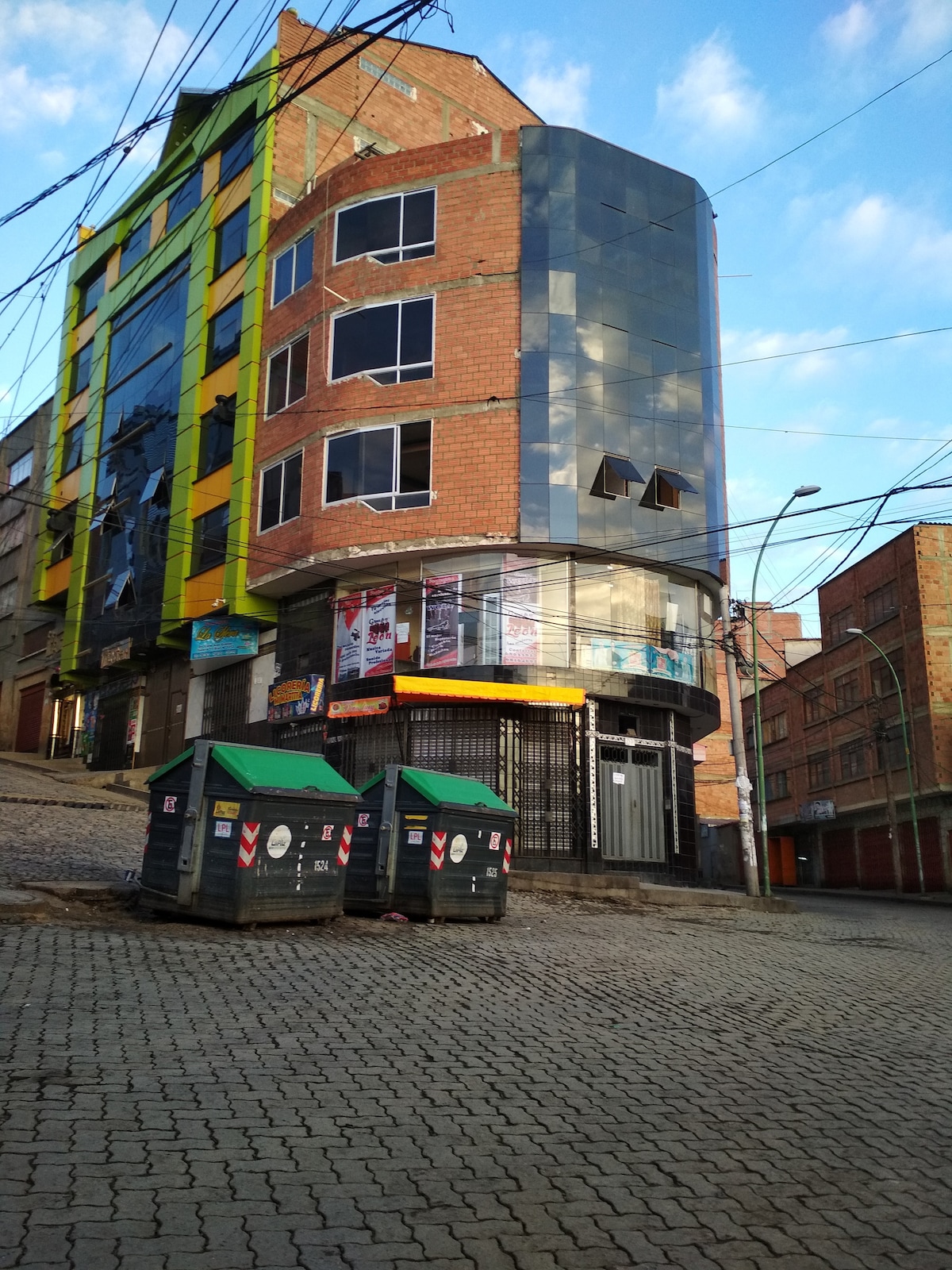 The image showcases a multi-story building with a modern design, featuring large glass windows and a curved facade. The ground level includes a storefront, while green dumpsters are visible in front. The setting is characterized by a sloped street, with power lines overhead and blue skies in the background.