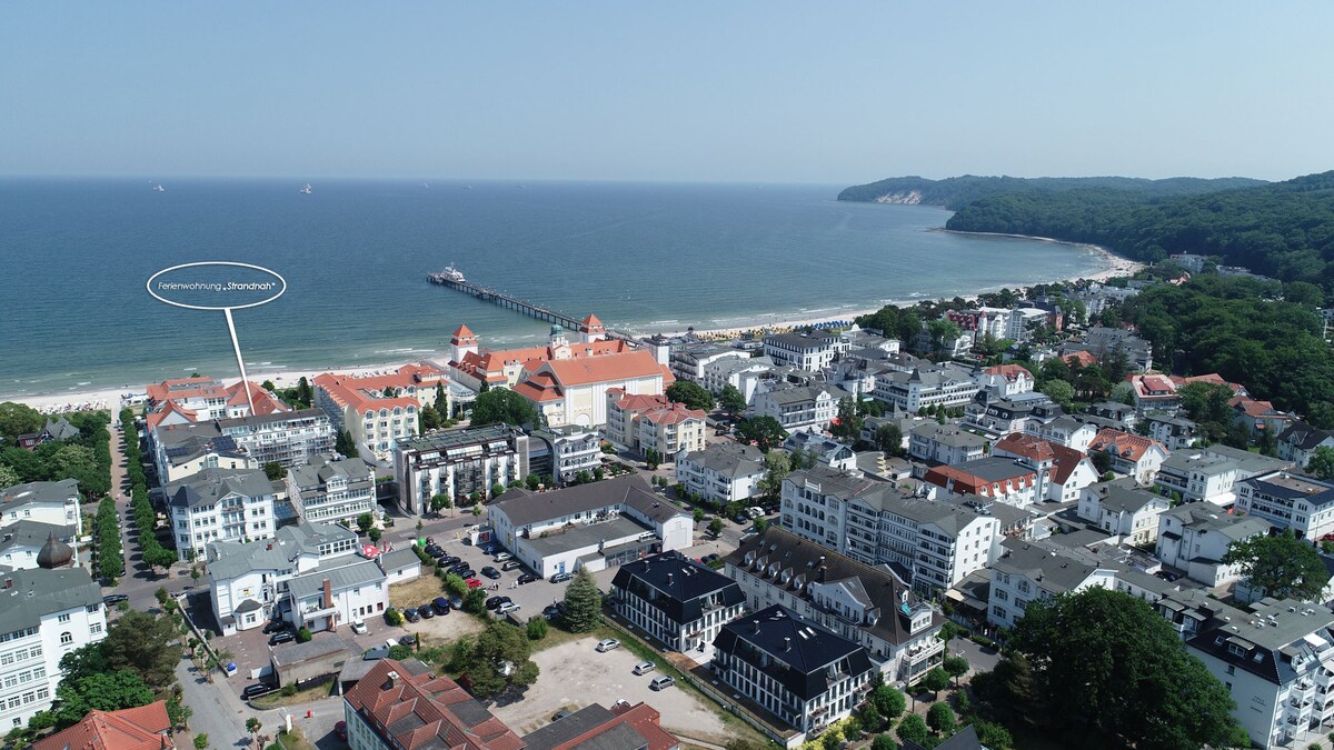 An aerial view captures the coastal area, highlighting the proximity of buildings to the beach. The shoreline is visible alongside the pier extending into the ocean. Nearby structures showcase a mix of architectural styles, with green trees offering a backdrop to the vibrant seaside setting.