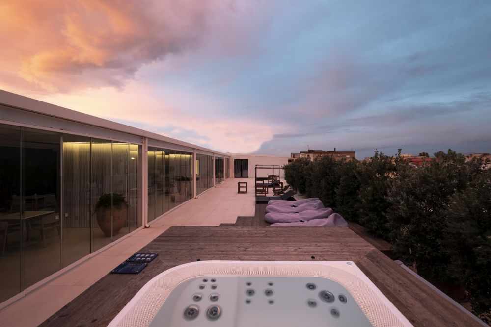 An outdoor area showcases a hot tub surrounded by wooden decking and soft lounge seating. The setting features modern architecture with large glass windows and a vibrant sky at sunset, creating a relaxing environment for social gatherings.