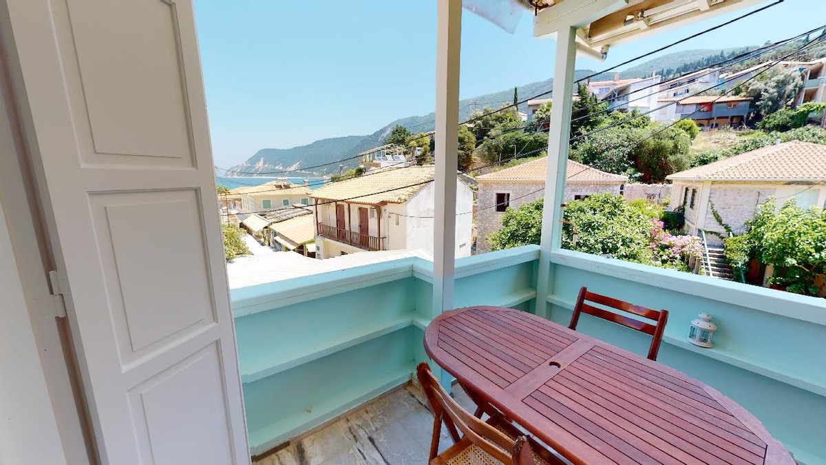 A balcony area is depicted, featuring a wooden table with matching chairs. Lush greenery and distant hills are visible, providing a scenic backdrop. Bright blue skies and sunlight enhance the open atmosphere of the space.