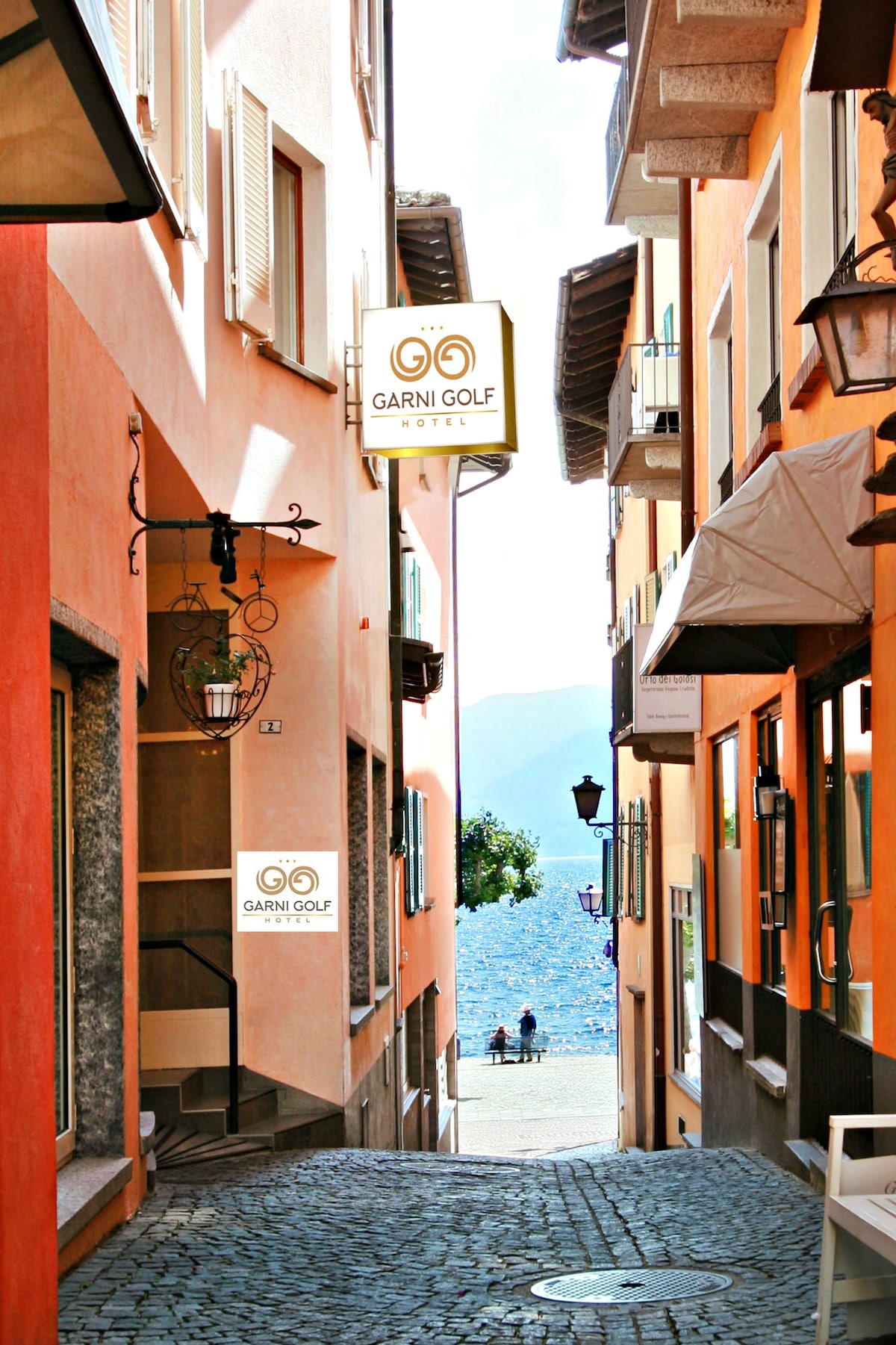 A charming alleyway lined with colorful buildings leads toward a view of the lake. Illuminated signs for the Garni Golf Hotel can be seen on either side, while cobblestone pavement creates a textured surface underfoot. The distant view features individuals sitting near the water.