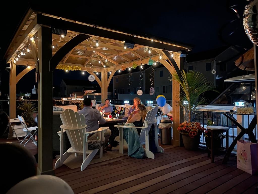 A spacious gazebo is illuminated by string lights, providing a cozy gathering space on a deck. Several people are seated around a table, enjoying conversation. Decorative elements and colorful balloons are arranged around the area, creating a festive atmosphere.