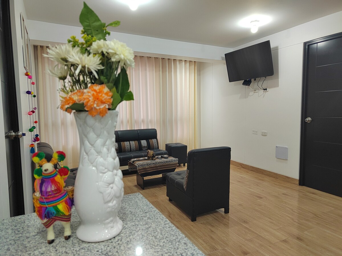 A cozy living area is presented with a dark sofa set and a vibrant decorative vase filled with flowers. Natural light enters through light-filtering beige curtains, and a television is mounted on the wall, creating a comfortable space for relaxation.