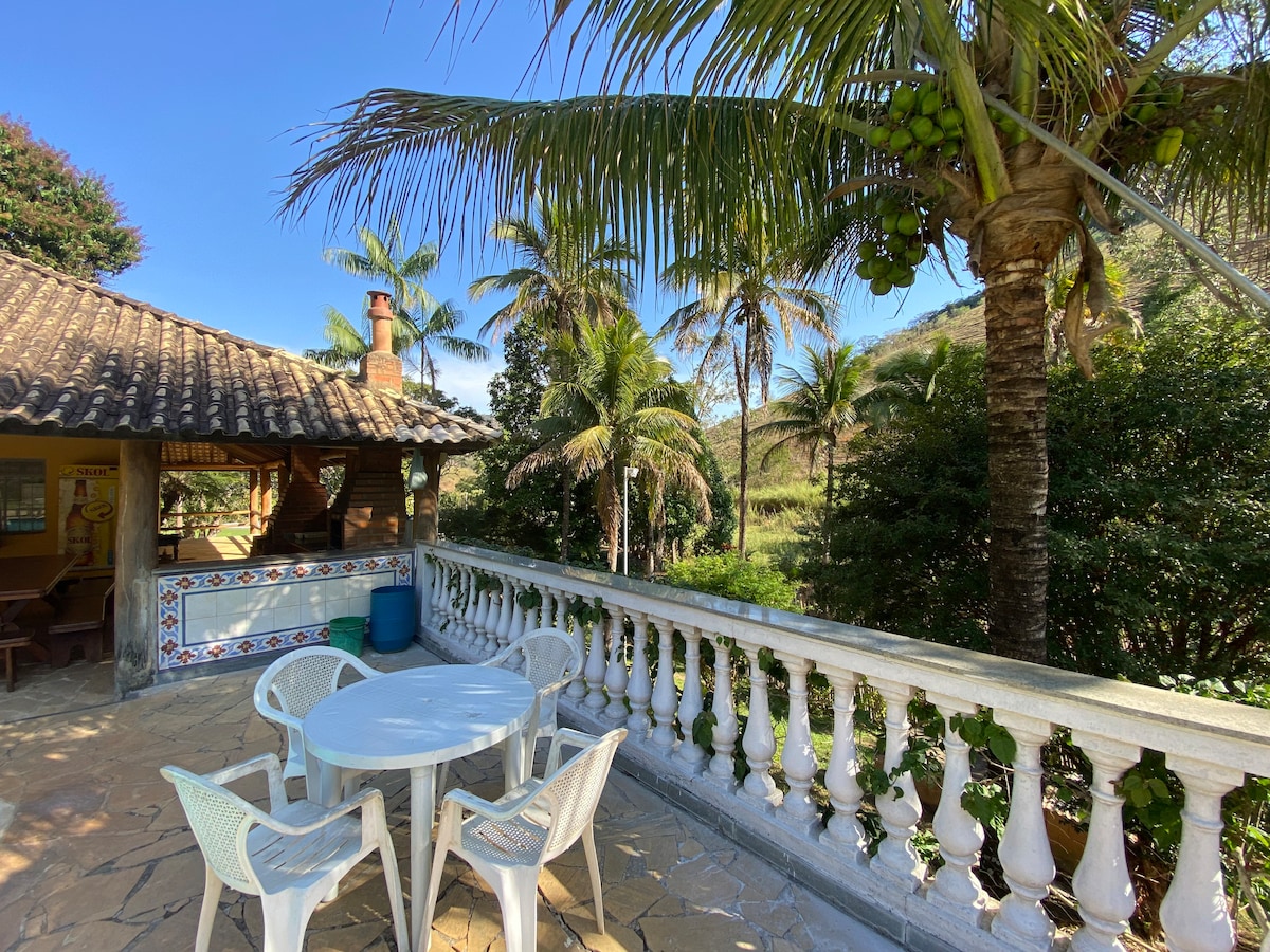 A balcony area is showcased with a round table surrounded by four white chairs. Lush palm trees are seen in the background, enhancing the natural setting. The rustic structure features a tiled roof and traditional decor, creating a serene outdoor space.