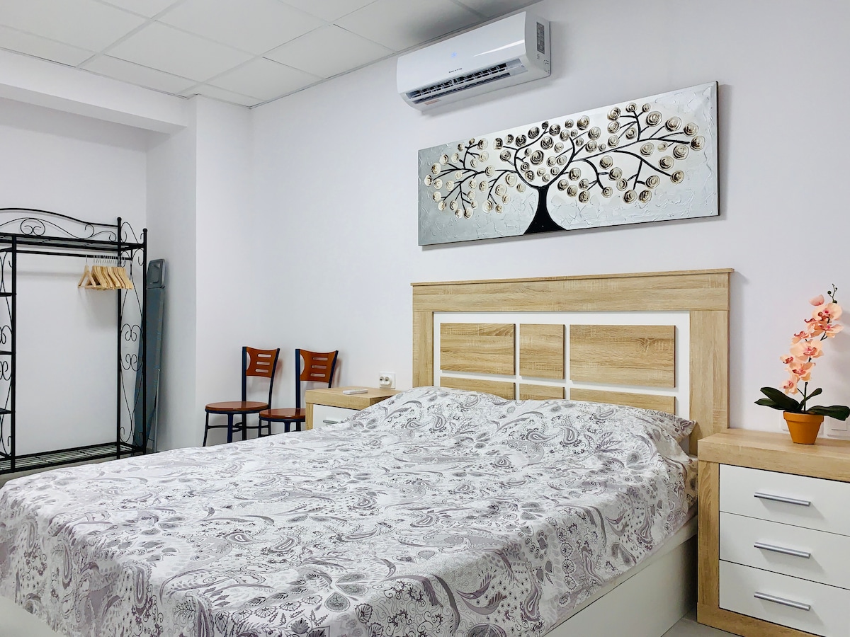 A bedroom features a spacious bed covered with a patterned comforter. A decorative wall piece depicting a tree is displayed above the headboard. Two wooden chairs are positioned nearby, and an air conditioning unit is mounted on the wall for climate control.