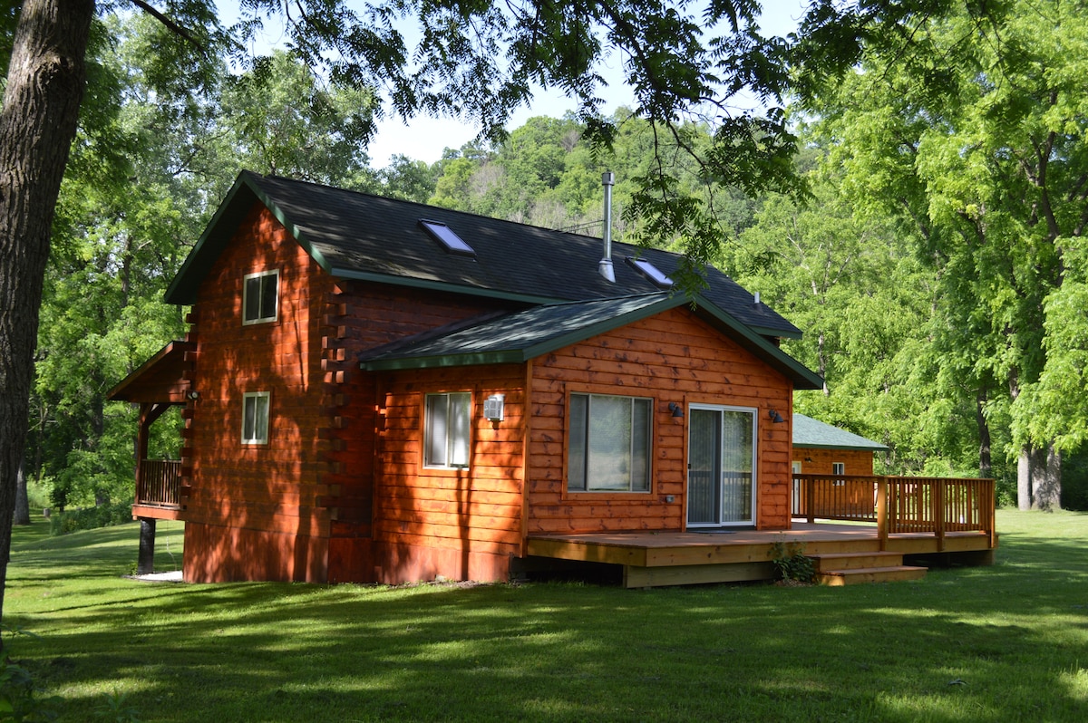 The exterior of a cabin is shown, featuring warm wooden siding and a large deck. Multiple windows allow natural light to fill the interior. The surrounding grassy area is complemented by lush green trees, creating a serene and inviting outdoor environment.