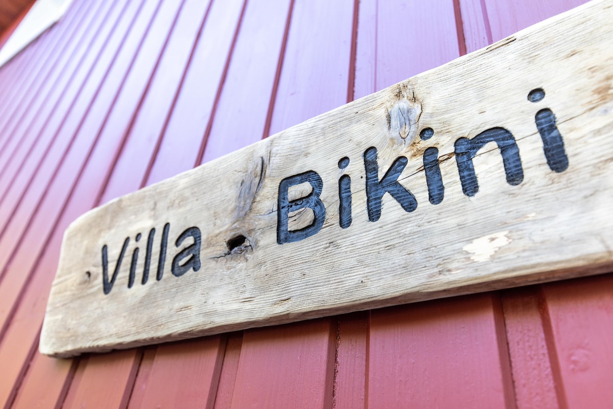 A wooden sign displaying the name 'Villa Bikini' is mounted on a red vertical wooden wall, showcasing a rustic touch. The sign features carved letters, indicating the villa's identity in a simple yet inviting manner.