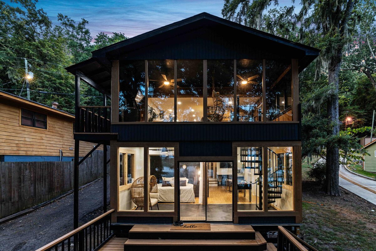 The exterior of a modern two-story house showcases large glass windows that provide expansive views of the surroundings. A spacious deck leads to the entrance, and a stylish spiral staircase is visible inside. The house is nestled within a natural setting, surrounded by greenery.
