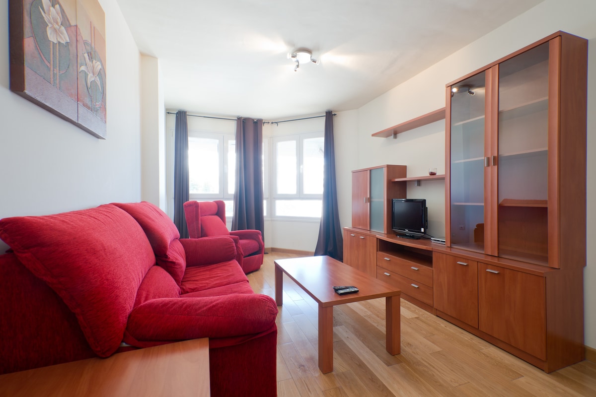 A living area is furnished with a red sofa set and a wooden coffee table. Natural light enters through large windows, framed by dark curtains. A wooden entertainment unit with a television sits against the wall, providing a cozy space for relaxation.