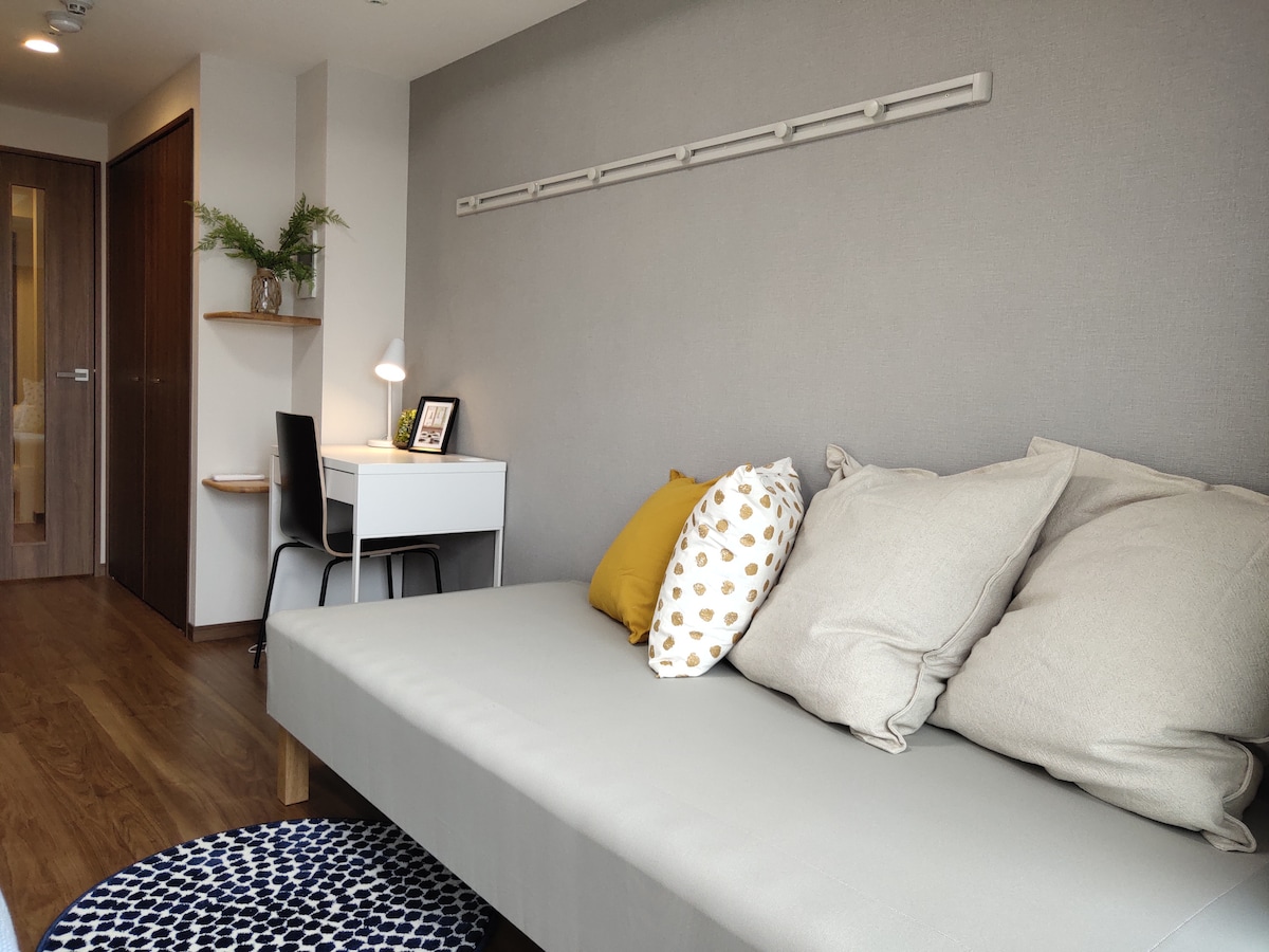 A light-filled room features a spacious sofa, adorned with several cushions and a cozy throw. A small desk with a lamp and a decorative picture frame is positioned against the wall. Light wood flooring contrasts with neutral wall tones.