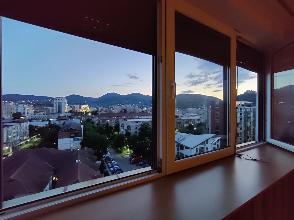A panoramic view is visible through large windows, offering glimpses of the surrounding city and rolling hills at dusk. The soft hues of the evening sky are reflected, while buildings are illuminated below, creating a serene urban landscape.