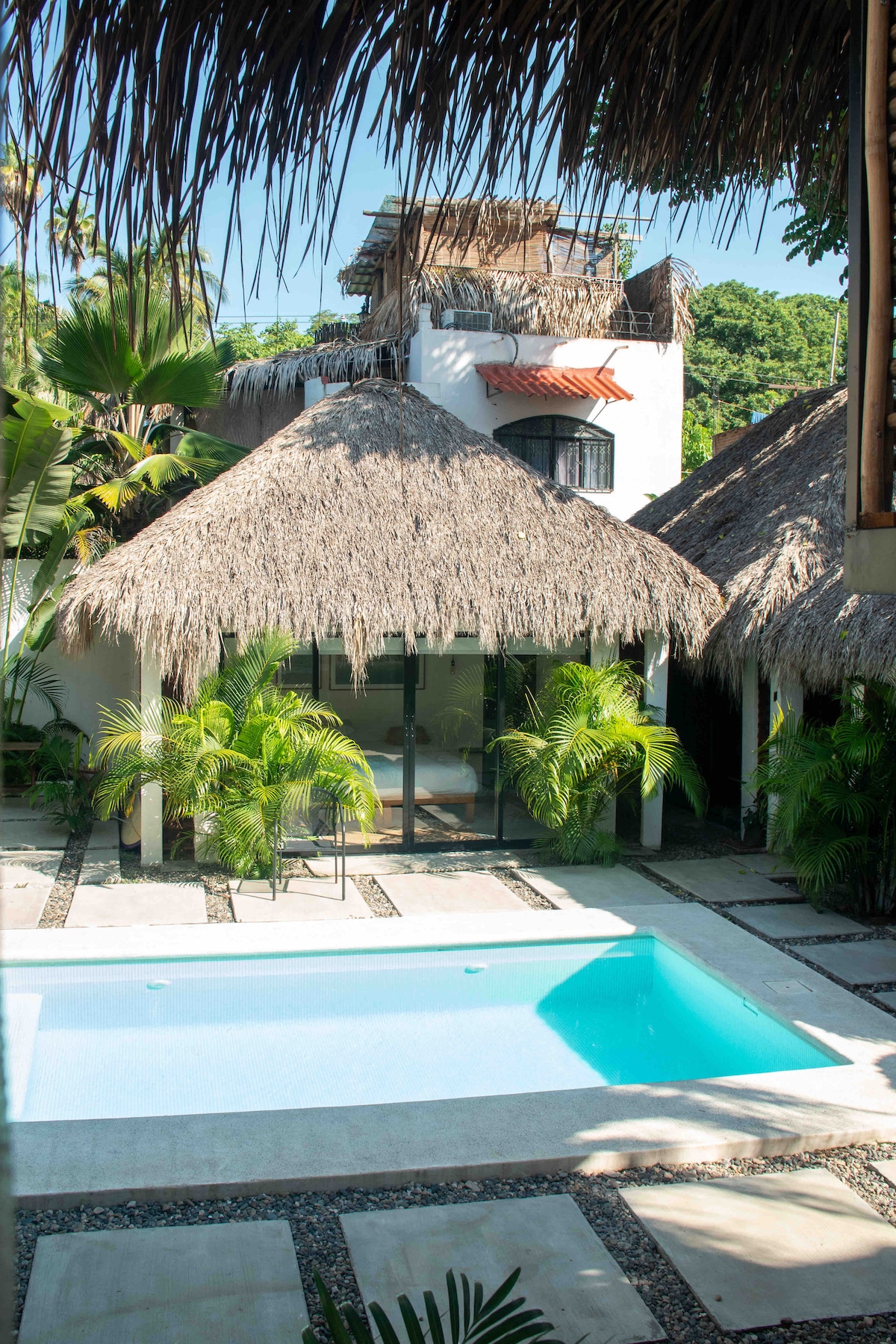 The image captures a shared pool area surrounded by thatched-roof bungalows. Lush green palm plants frame the pool, and the outdoor space features a stone pathway leading to a tranquil swimming area. Sunlight illuminates the scene, enhancing the serene atmosphere.