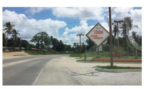 A welcoming sign is positioned along a clear road, indicating the entrance to a community. Lush palm trees and scattered greenery line the street, with a partly cloudy sky providing bright, natural light.