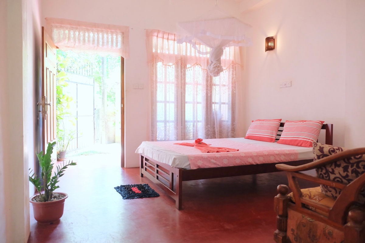 A spacious room is featured with a large bed adorned with soft linen. Natural light filters through sheer curtains covering multiple windows. A potted plant adds a touch of greenery, while a comfortable chair complements the space. The entrance leads to an outdoor area.