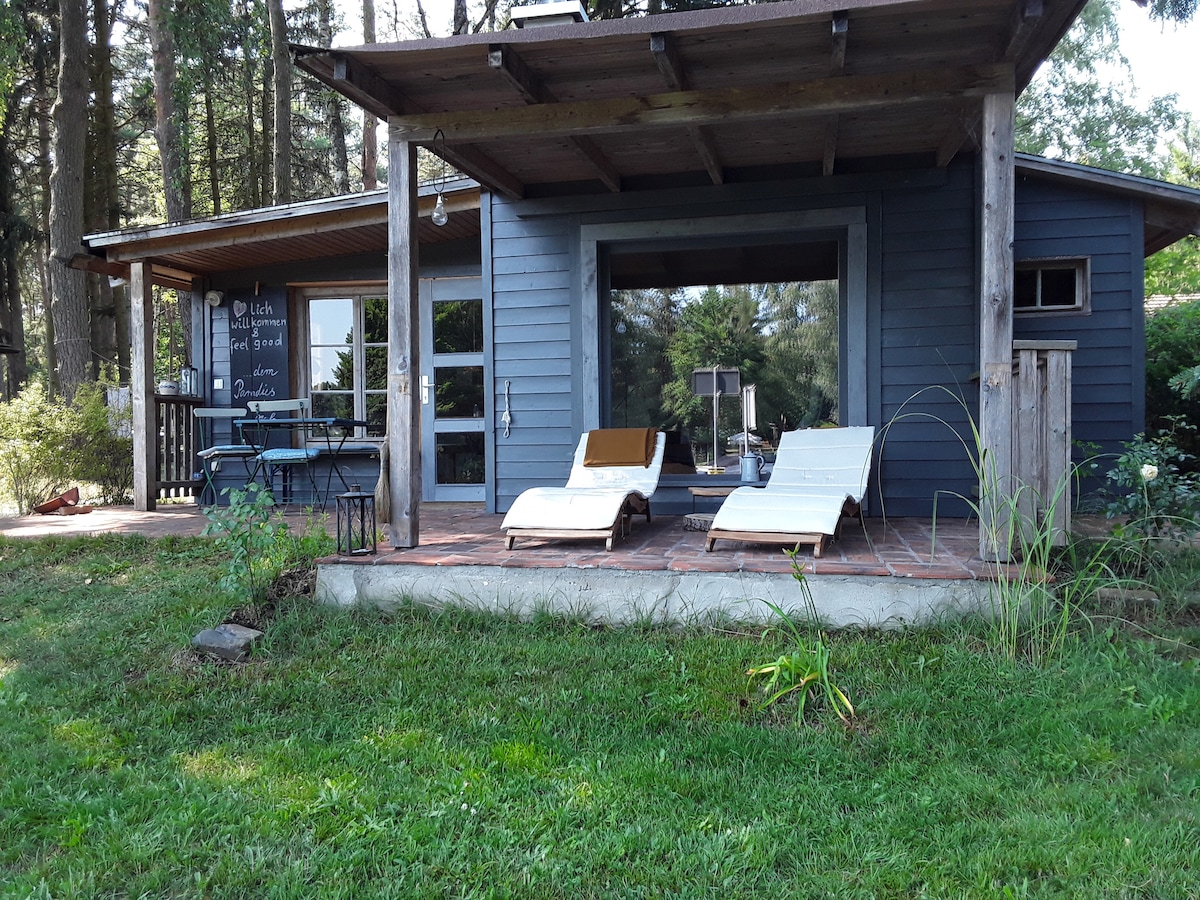 A welcoming cabin is set amidst lush greenery, featuring a covered patio with two lounge chairs. Large windows provide views of the forest, while a small dining area is visible on the adjacent terrace. The exterior combines natural wood elements with modern design.