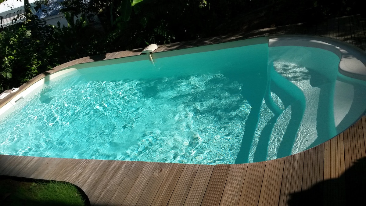 A clear swimming pool is set within a wooden deck, reflecting sunlight and surrounded by lush greenery. The inviting water features gentle ripples, creating a serene ambiance. Part of the pool is shaded, providing a cool area for relaxation.