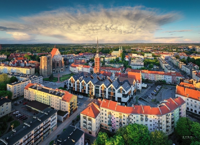 An aerial view captures a vibrant cityscape featuring a mix of modern buildings and historical architecture. Surrounding greenery adds a touch of nature, while a dramatic sky filled with clouds creates a striking backdrop over the neighborhood, highlighting the urban layout.