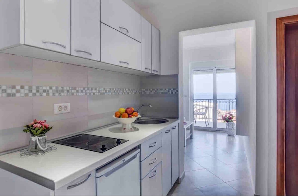 A modern kitchen with white cabinetry features a fruit bowl on the countertop. Nearby, a sink and stove are positioned for convenience. A doorway leads to a bright area with a view of the sea, accented by natural light and floral arrangements.