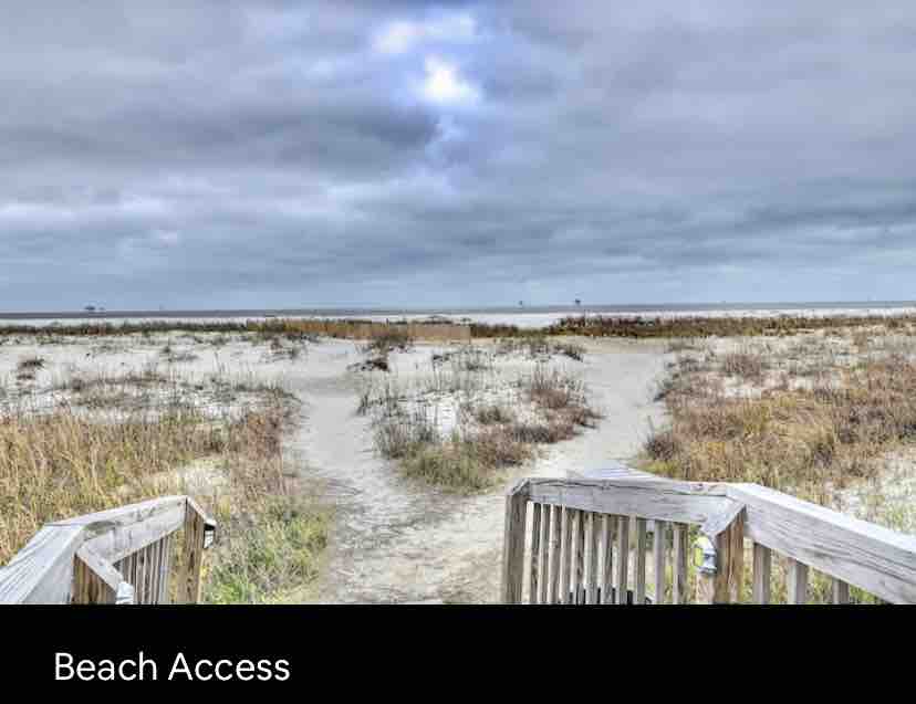 A wooden boardwalk leads to the beach, surrounded by natural sand and coastal vegetation. The open sky presents a blend of gray clouds, suggesting an overcast day, while the distant horizon meets the water of the Gulf.