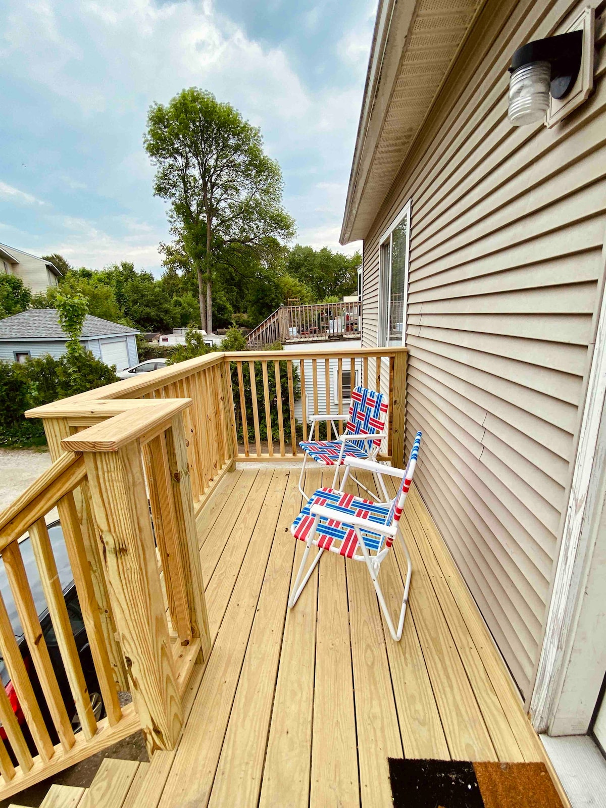 A small wooden deck is featured, showcasing two striped folding chairs arranged for relaxation. The deck is framed by wooden railings and extends from the side of the building, overlooking a green landscape with trees and nearby parking.