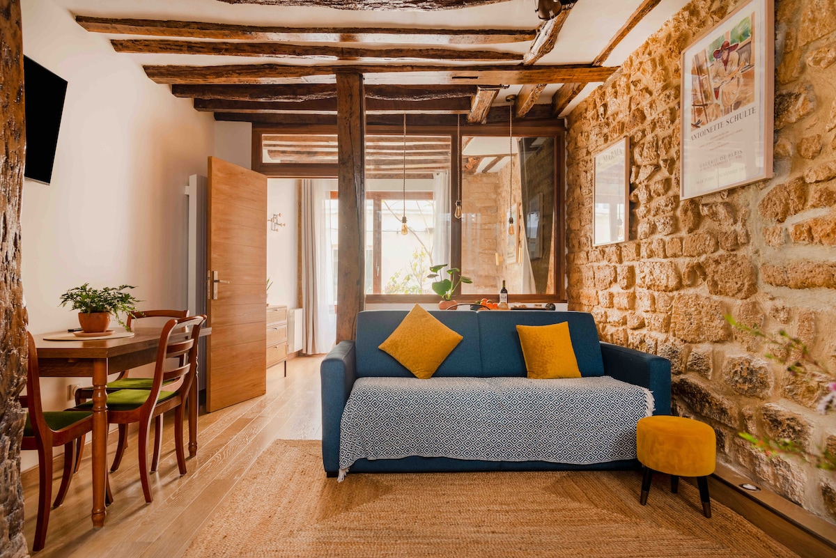 A cozy living area features a blue sofa adorned with a mustard yellow cushion. A small round ottoman sits nearby. Light wooden floors complement exposed stone walls and rustic wooden beams. A dining table for four is positioned to the left, with greenery adding a touch of warmth.