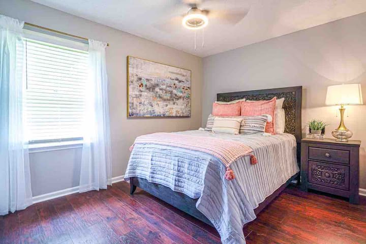 سرير مريح بحجم كوين مع لحاف وملاءات رمادية ناعمة خفيفة الوزن. منضدة جانبية واسعة مع مصباح نحاسي. مروحة سقف حديثة وصورة تجريدية كبيرة وجميلة على الجدار. 