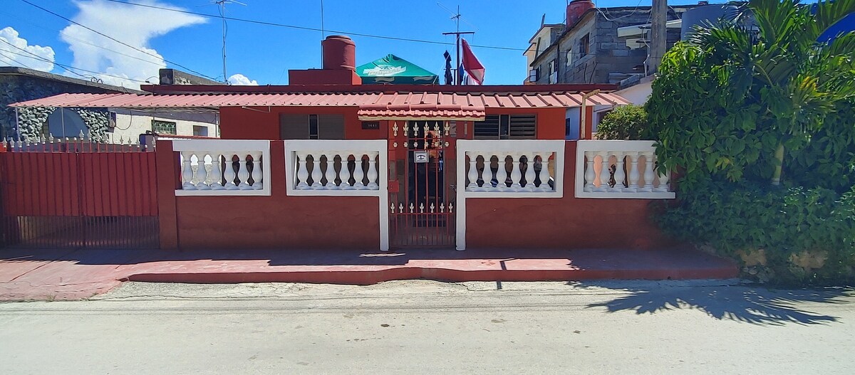 The exterior of a red-colored building is framed by a white wall with decorative balusters. A gated entrance leads to a pathway, flanked by tropical greenery. A shaded area with a green umbrella is visible above the main entrance.