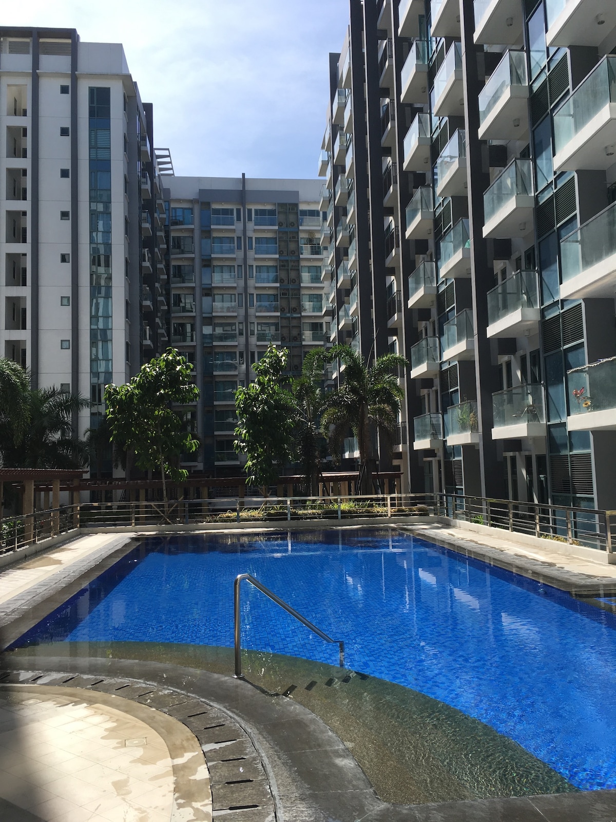 A clean swimming pool is surrounded by modern apartment buildings. Lush green trees and palm plants are positioned near the pool area, providing a blend of nature amidst the urban setting. Bright sunlight reflects off the water, creating a serene atmosphere.