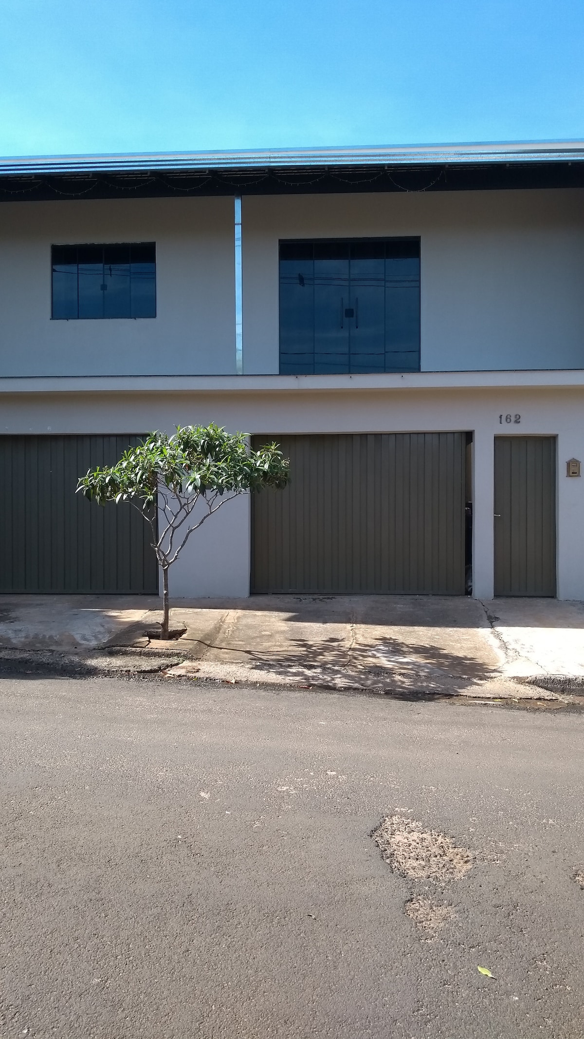 A two-story structure with a light-colored exterior is visible. The building features two large windows on the upper floor and two garage doors on the ground floor. A small tree is positioned in front of the entrance, providing a touch of greenery to the scene.