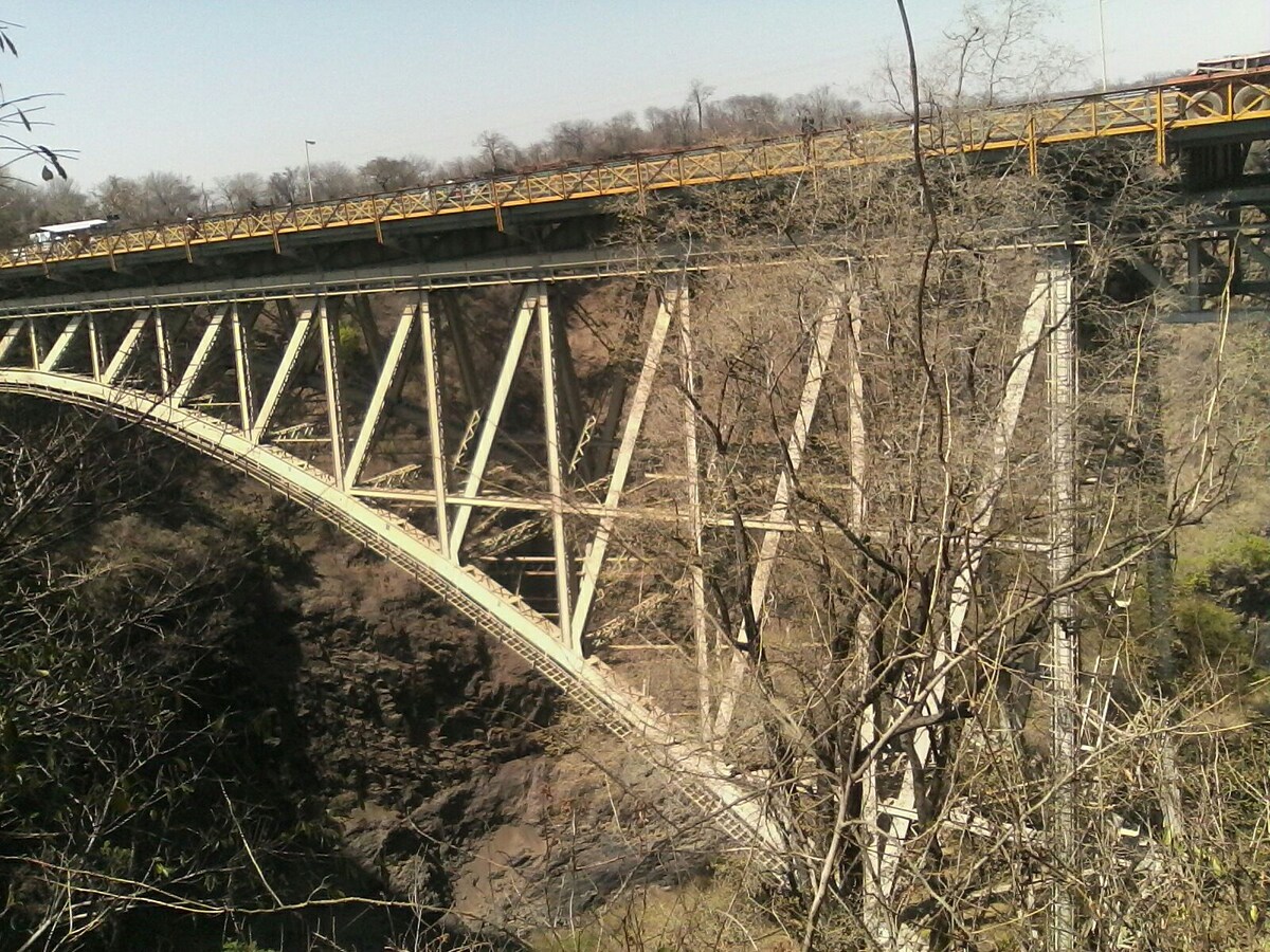 A large bridge is prominently depicted, showcasing a strong metal framework above a deep gorge. Surrounding vegetation includes sparse trees and brush, emphasizing the rugged natural terrain. The clear sky above creates a sense of open space in the image.
