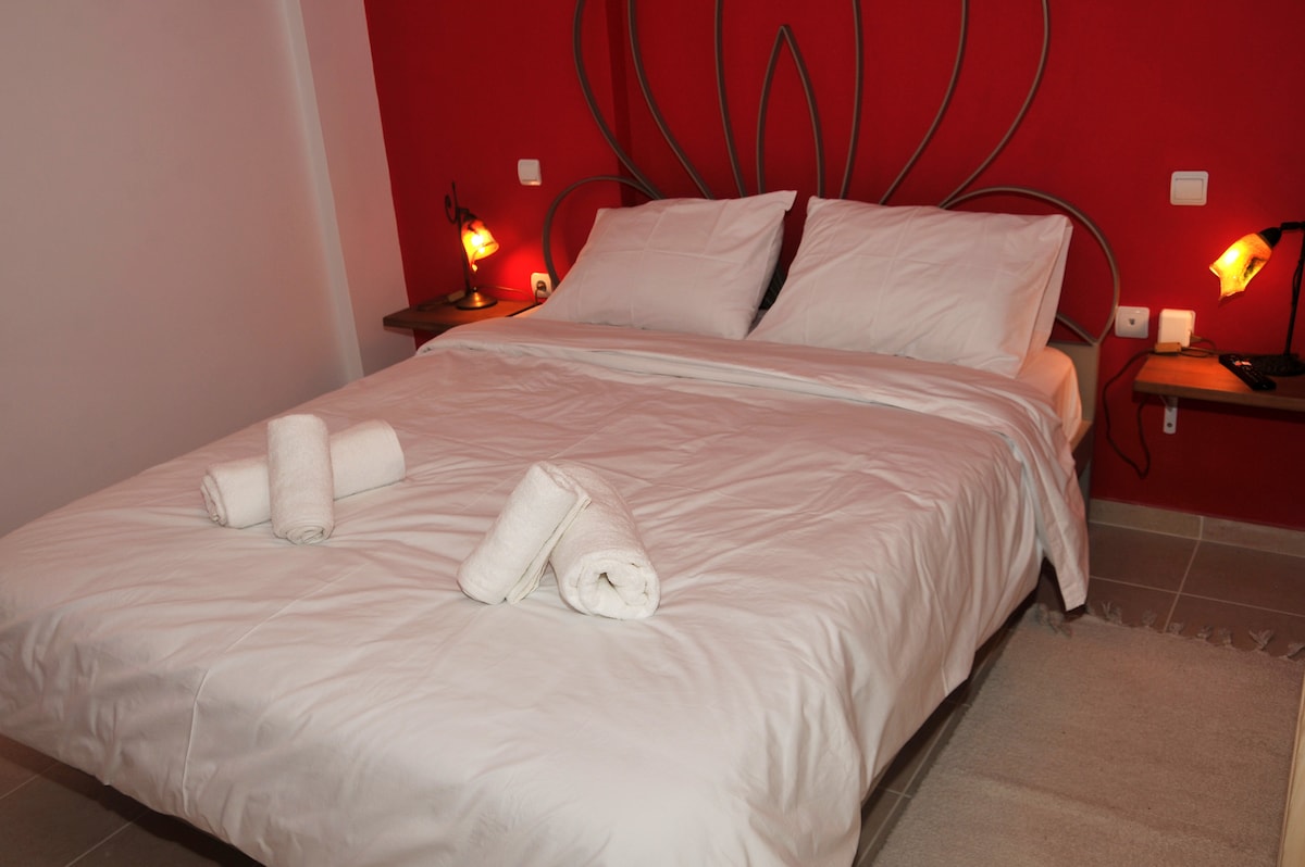 A spacious bed is centered against a vibrant red wall, with decorative lamps on either side providing soft illumination. Fresh towels are neatly arranged on the bed, adding a touch of cleanliness to the inviting environment.