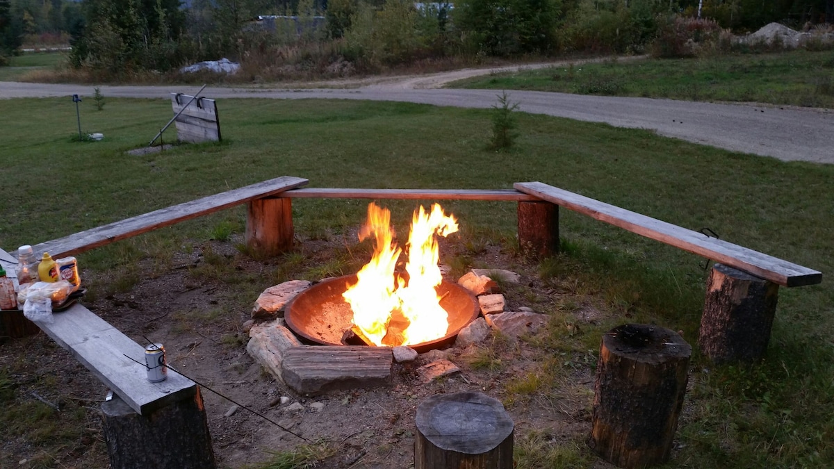 A fire pit is surrounded by rustic wooden benches, offering a cozy gathering space. The fire is brightly glowing, casting light on the surrounding grassy area. In the background, a gravel road is visible, bordered by trees and natural landscape.