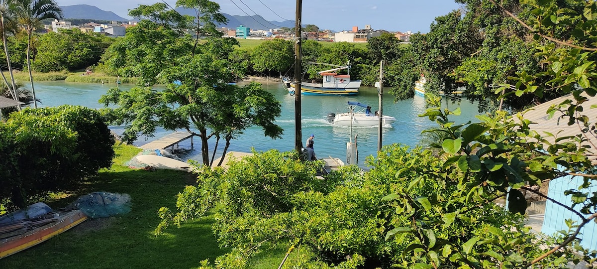 The image captures a serene canal scene, framed by lush greenery. Two boats are seen navigating the water, while a small dock with a fisherman is visible. The surrounding area features a mix of residential buildings and nature, contributing to a calm ambiance.