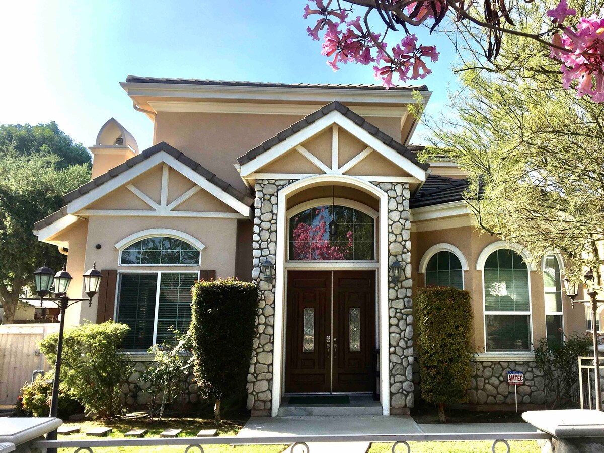 The exterior of a charming home is showcased, featuring a combination of stone and stucco finishes. Large windows allow natural light to enter, while a stately double door entrance is framed by greenery and flowering plants. Decorative lanterns provide an inviting touch.
