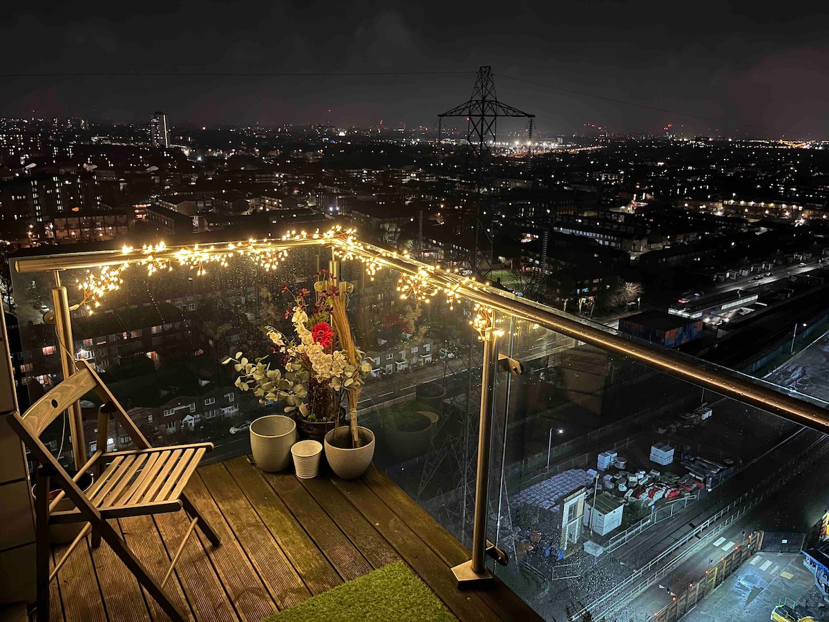 A balcony is illuminated with string lights, overlooking the sprawling cityscape at night. Potted plants and flowers add a touch of nature to the scene, while the distant lights of London create a vibrant backdrop.