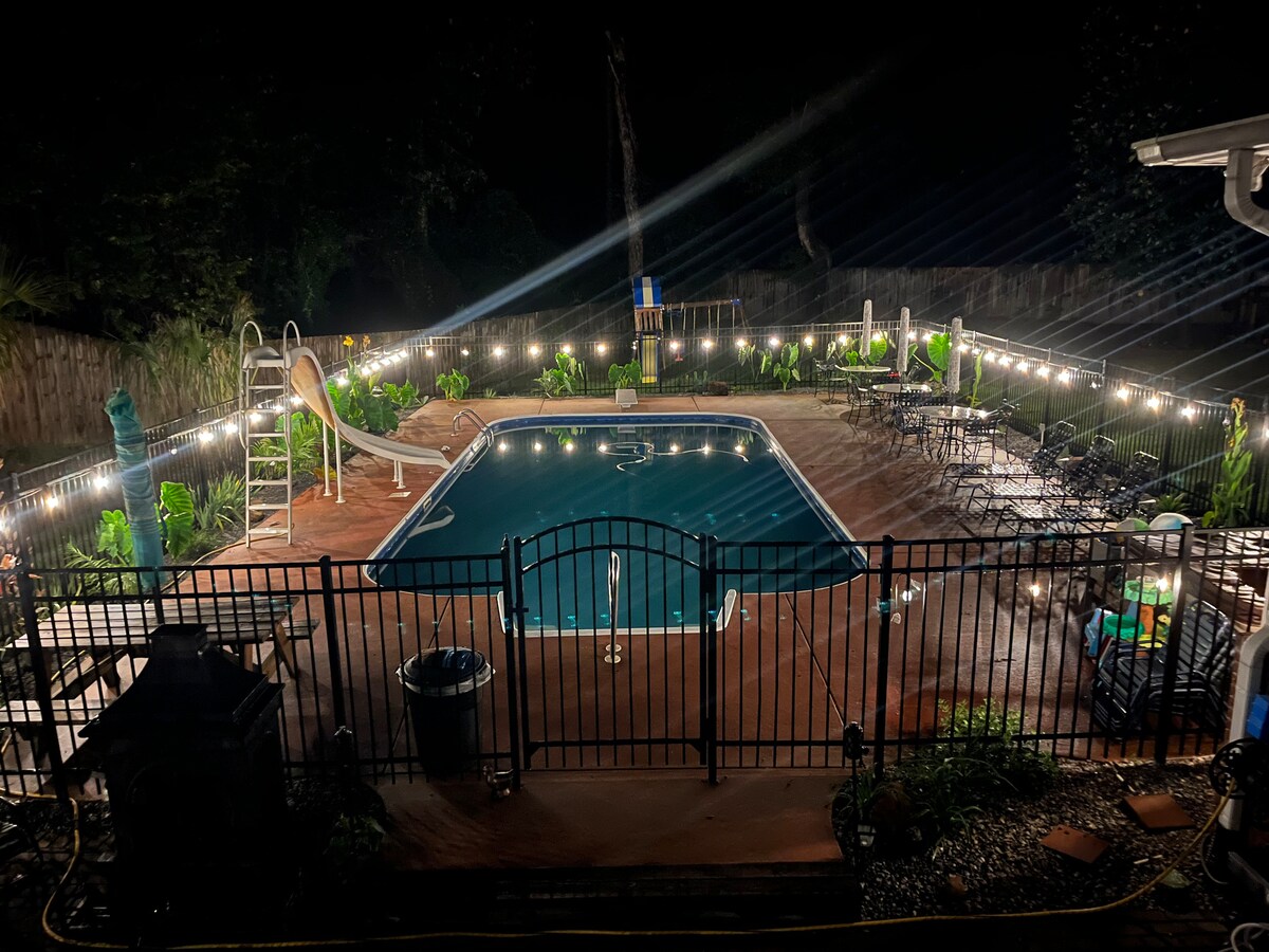 The in-ground pool is surrounded by a secure fence and features lounge seating and a waterslide. String lights illuminate the area, creating a gentle glow, while landscaped greenery is visible around the pool deck, enhancing a serene outdoor setting.