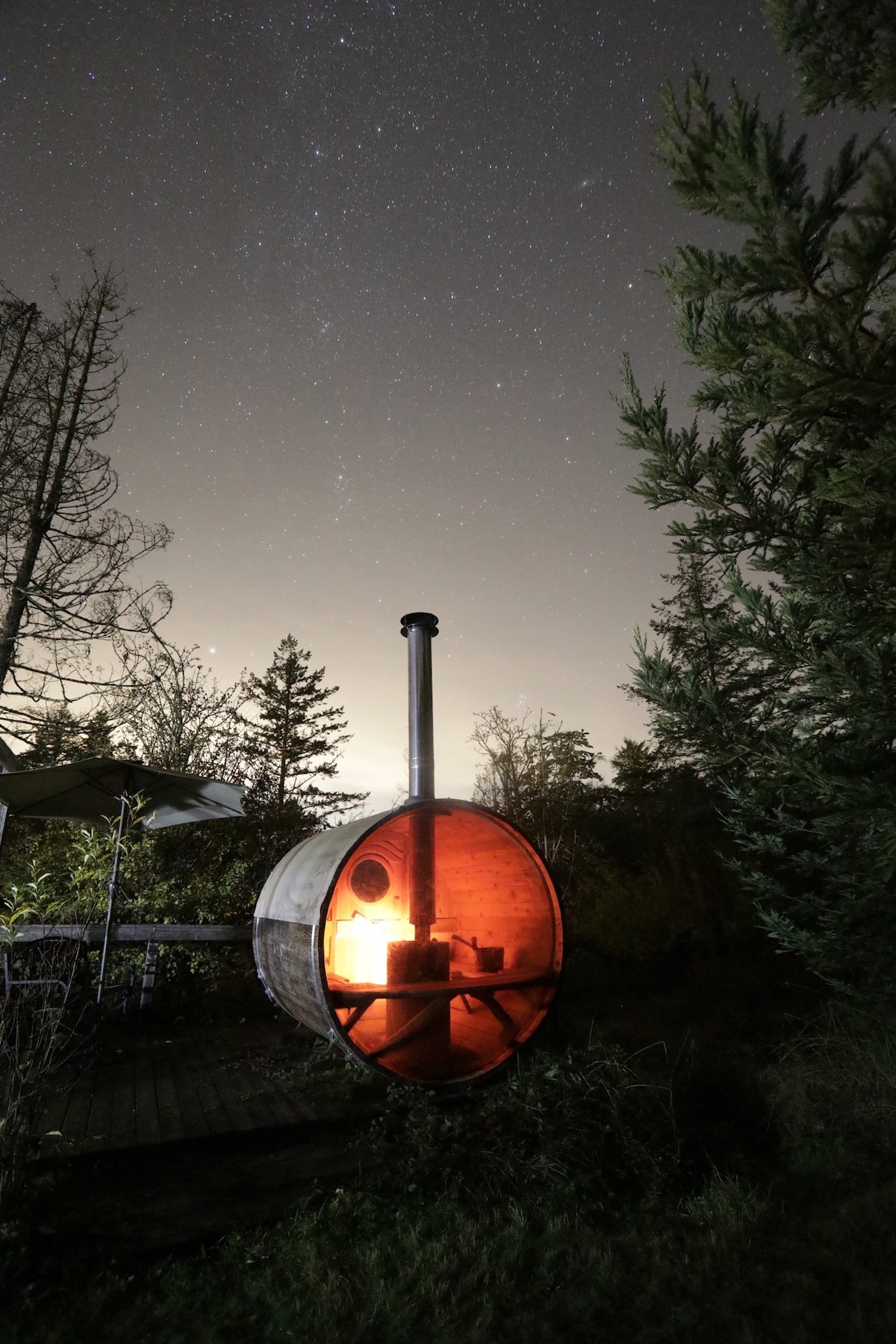 A wood-burning barrel sauna is illuminated softly by its interior glow under a starry night sky. Nearby trees frame the scene, while the surrounding landscape is bathed in a gentle twilight. The structure is positioned on a wooden deck, inviting relaxation in a serene setting.