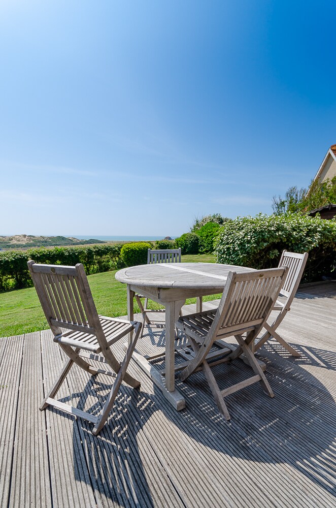 An outdoor dining area features a round table surrounded by six wooden chairs. The area is set on a deck, with lush green grass and hedges visible. A clear blue sky provides a bright backdrop, enhancing the inviting outdoor space.