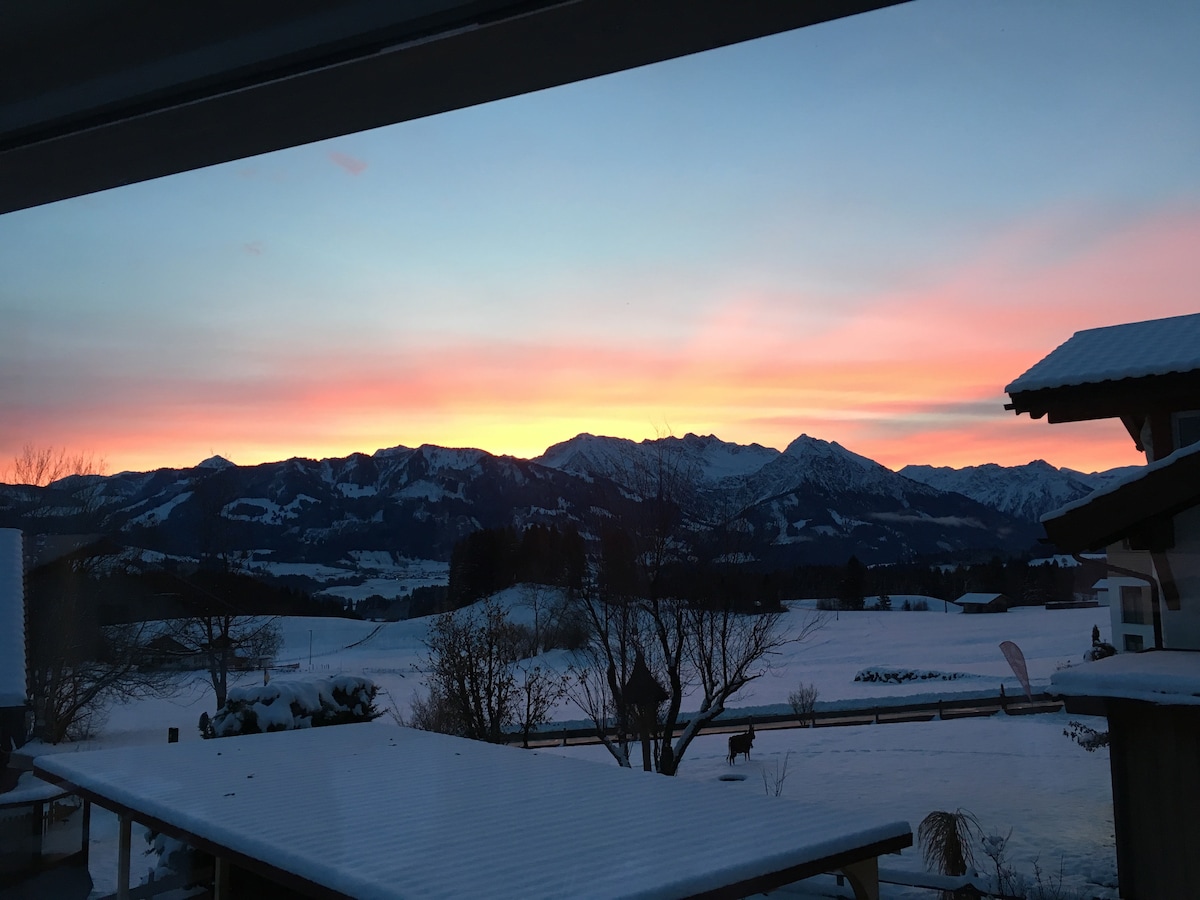 A stunning winter landscape is captured at dusk, featuring snow-covered ground and gently rolling hills. The sky displays a gradient of colors from soft pink to deep orange, framing the silhouette of distant mountains. A tree stands prominently in the foreground.