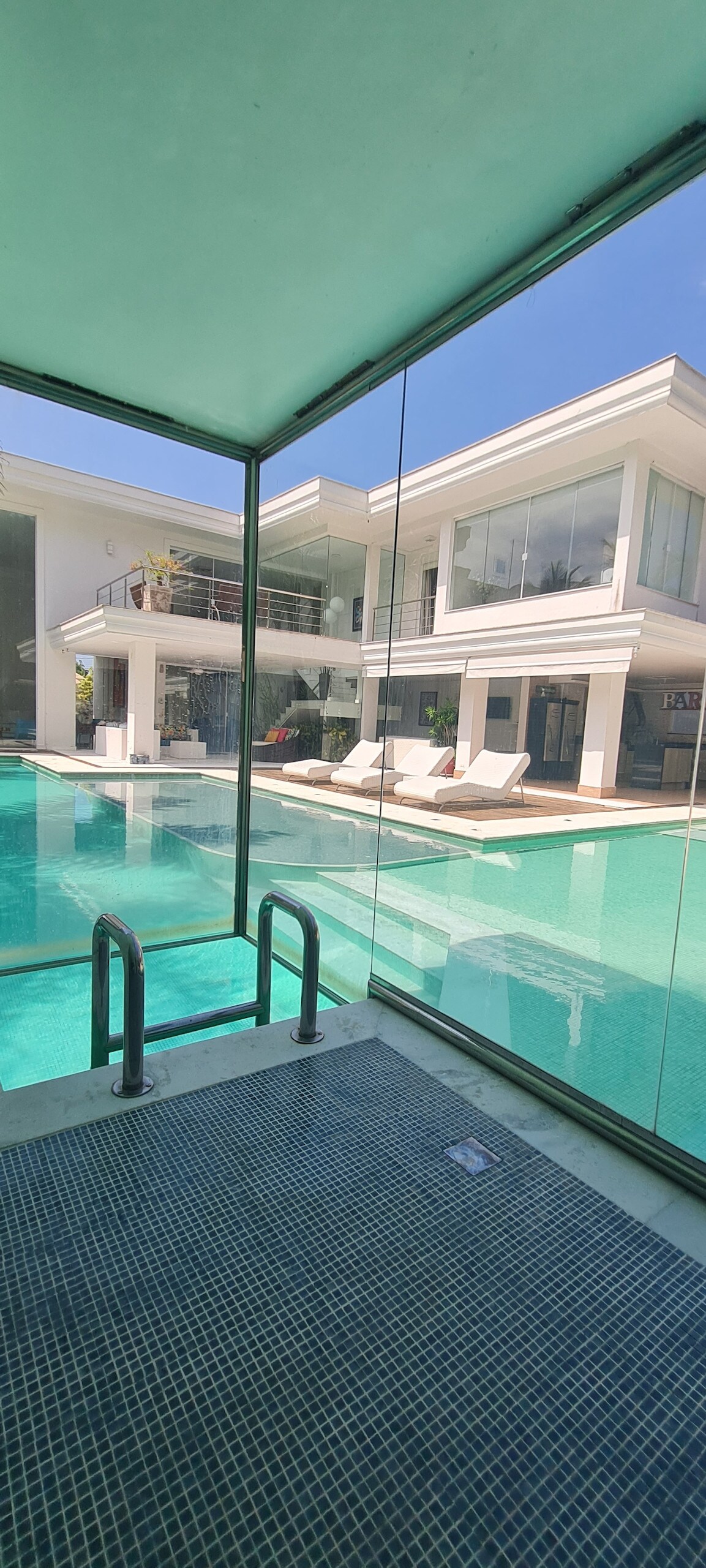 The view from an indoor lounge area showcases a spacious pool surrounded by modern lounge chairs. Large glass walls provide a clear sight of the two-story house's exterior, reflecting sunlight and the serene pool waters.