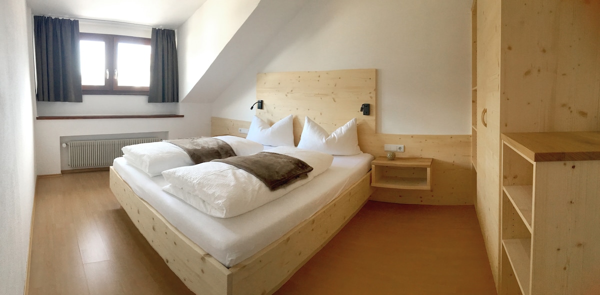 A spacious sleeping area features a wooden bed with two large pillows and two decorative cushions. Natural light enters through the windows, which are flanked by dark curtains. A shelving unit and bedside table complement the serene atmosphere.