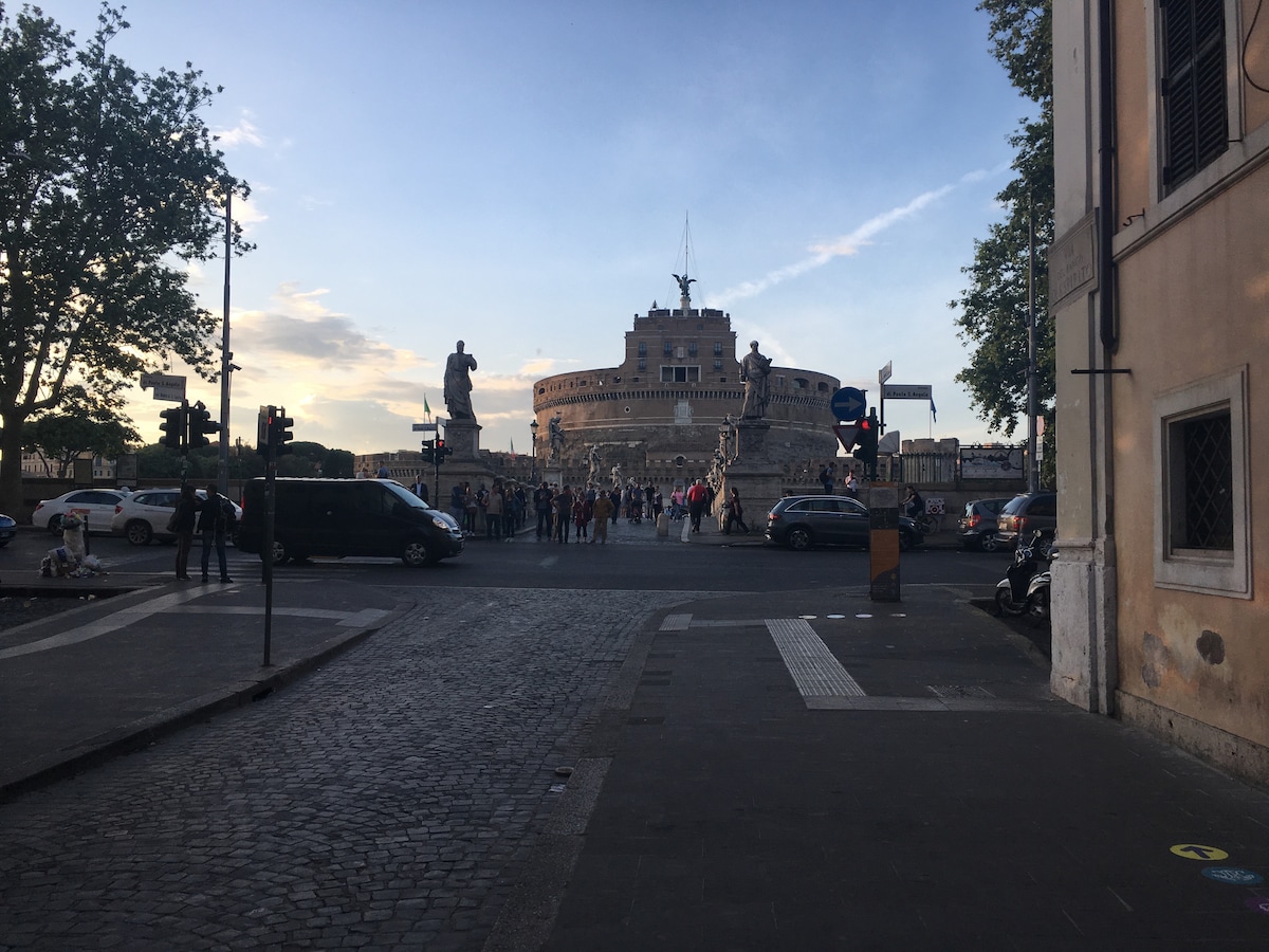 A lively street scene is presented, showcasing the historic Castel Sant'Angelo in the background. Vehicles and pedestrians are observed near a traffic light, with trees lining the road. A statue stands prominently at the intersection, adding to the area's character.